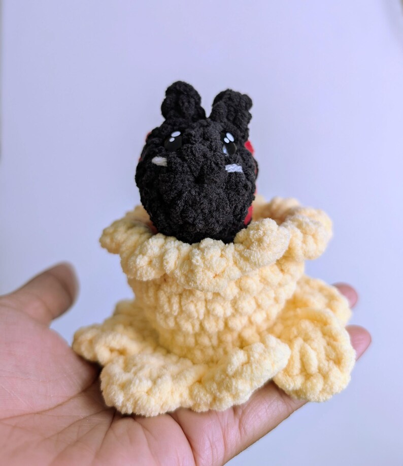 May include: A handmade crochet bunny with a black head and white eyes, nestled in a light yellow crochet flower. The bunny has red accents and is made of soft yarn.