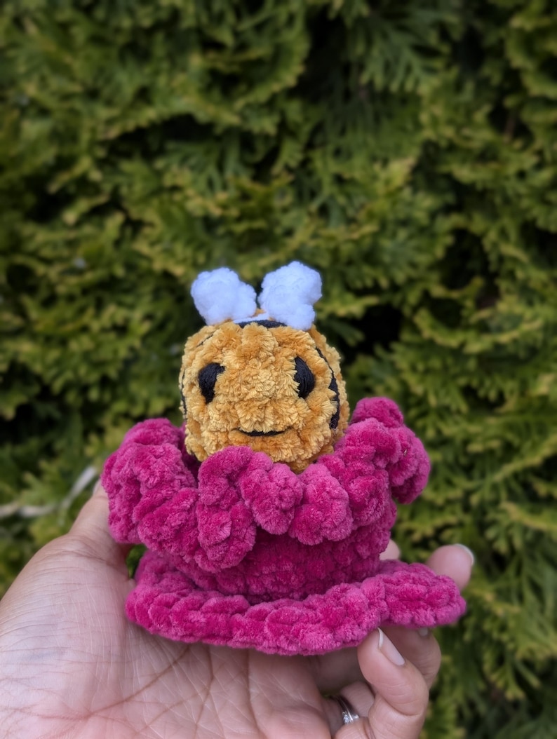 May include: A handmade crochet bee with a yellow and black body, white antennae, and a magenta flower-like base. The plush toy is held in a hand against a backdrop of green foliage.