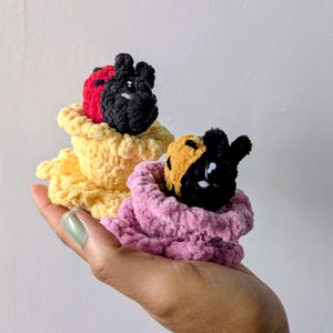 May include: Two handmade crochet creatures with black, red, and yellow color schemes. The creatures are nestled in yellow and pink crochet cups. The image is taken from a close-up angle, highlighting the texture and detail of the yarn.