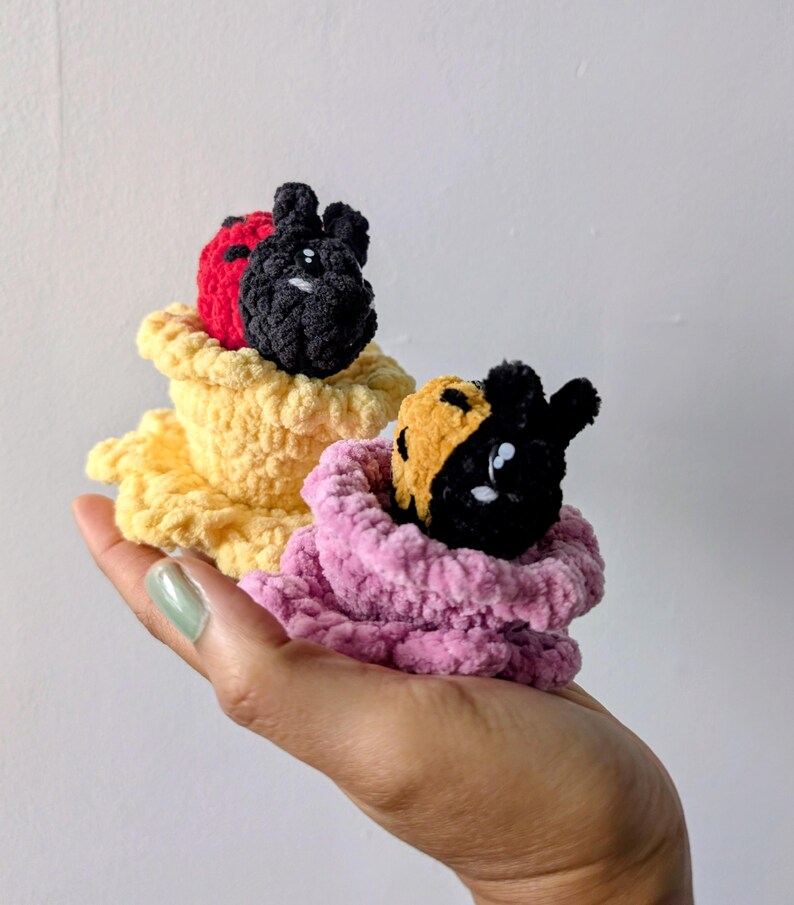 May include: Two handmade crochet creatures with black, red, and yellow color schemes. The creatures are nestled in yellow and pink crochet cups. The image is taken from a close-up angle, highlighting the texture and detail of the yarn.