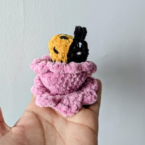 May include: A handmade crochet plush toy featuring a split-faced design: one half is yellow with black spots, the other is black. It sits in a pink, ruffled, plush base. The toy is held in a hand against a plain white background.