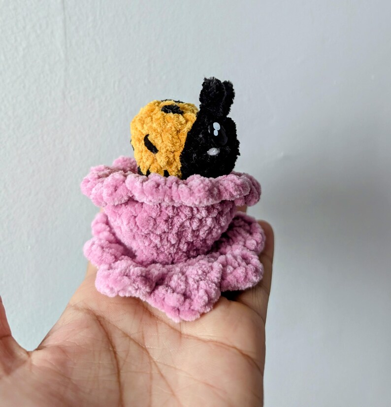 May include: A handmade crochet plush toy featuring a split-faced design: one half is yellow with black spots, the other is black. It sits in a pink, ruffled, plush base. The toy is held in a hand against a plain white background.