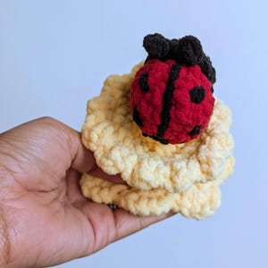 May include: A handmade crochet ladybug with a red body, black spots and stripes, and black antennae, atop a yellow, ruffled crochet base. The ladybug is a whimsical, soft toy.