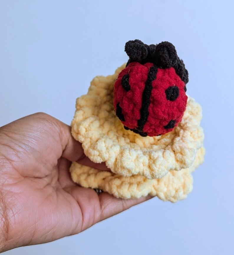 May include: A handmade crochet ladybug with a red body, black spots and stripes, and black antennae, atop a yellow, ruffled crochet base. The ladybug is a whimsical, soft toy.
