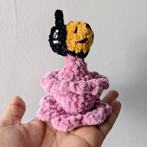 May include: A handmade plush toy with a black and yellow face and a pink, tiered body. The face has a smiling expression and a single eye. The toy is made of soft, textured yarn and is held in a hand.