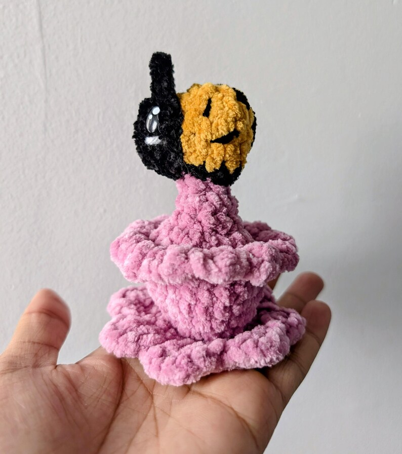 May include: A handmade plush toy with a black and yellow face and a pink, tiered body. The face has a smiling expression and a single eye. The toy is made of soft, textured yarn and is held in a hand.