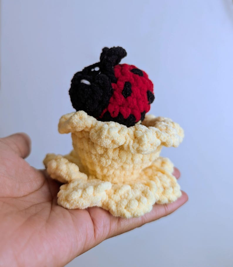 May include: A handmade crochet ladybug with a red and black body, perched in a light yellow crochet flower. The ladybug has black spots and a black head. The flower has a ruffled edge.