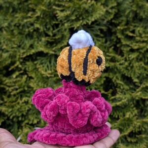 May include: A handmade crochet bee with a yellow and black striped body and white top, perched atop a vibrant pink, ruffled base. The plush toy is designed with soft, textured yarn, creating a whimsical and tactile appeal.