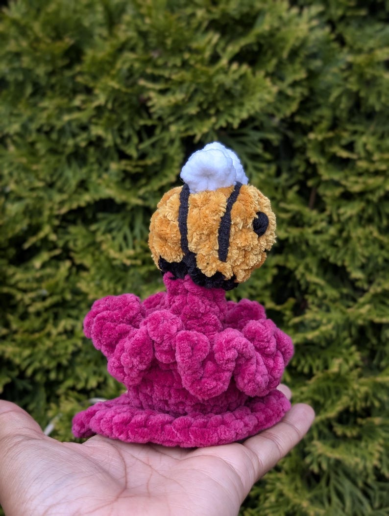 May include: A handmade crochet bee with a yellow and black striped body and white top, perched atop a vibrant pink, ruffled base. The plush toy is designed with soft, textured yarn, creating a whimsical and tactile appeal.