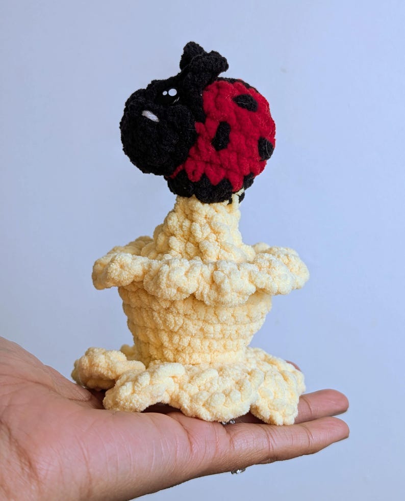 May include: A handmade crochet ladybug figurine with a red and black head and a cream-colored body. The ladybug has black spots on its red shell and is perched atop a ruffled, cream-colored base. The figurine is held in a hand.
