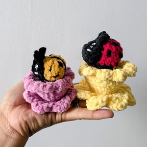 May include: Two handmade crochet plush toys: a bee and a ladybug. The bee has a black and yellow body, and the ladybug has a black and red body. Both are nestled in a pink and yellow flower-like base. The toys are held in a hand.