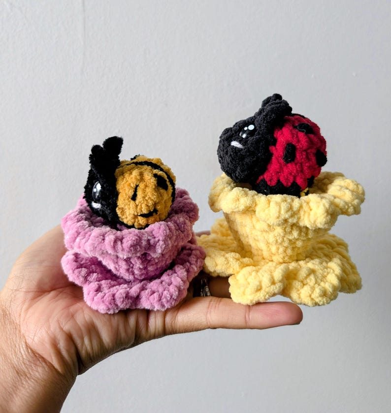 May include: Two handmade crochet plush toys: a bee and a ladybug. The bee has a black and yellow body, and the ladybug has a black and red body. Both are nestled in a pink and yellow flower-like base. The toys are held in a hand.
