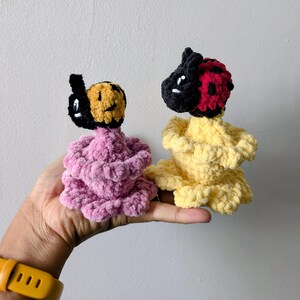 May include: Two handmade crochet dolls. One doll has a black and yellow head and a pink body. The other doll has a black and red head and a yellow body. The dolls are held in a hand against a white background.