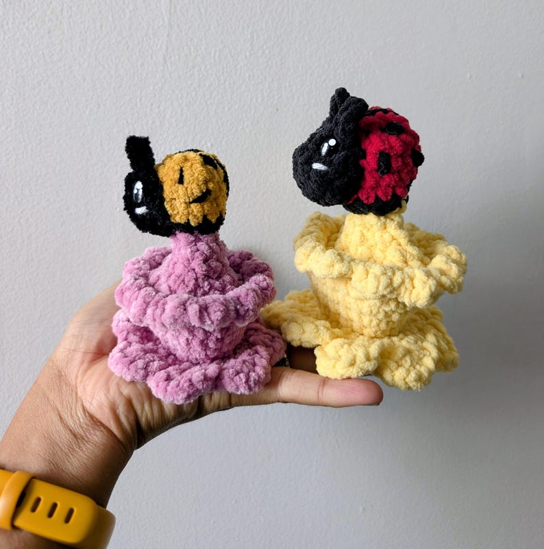 May include: Two handmade crochet dolls. One doll has a black and yellow head and a pink body. The other doll has a black and red head and a yellow body. The dolls are held in a hand against a white background.