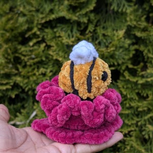 May include: A handmade plush bee with black stripes and a white head, perched atop a vibrant magenta flower. The bee is made of soft, textured material, and the flower has a ruffled appearance. The background is a blurred green.