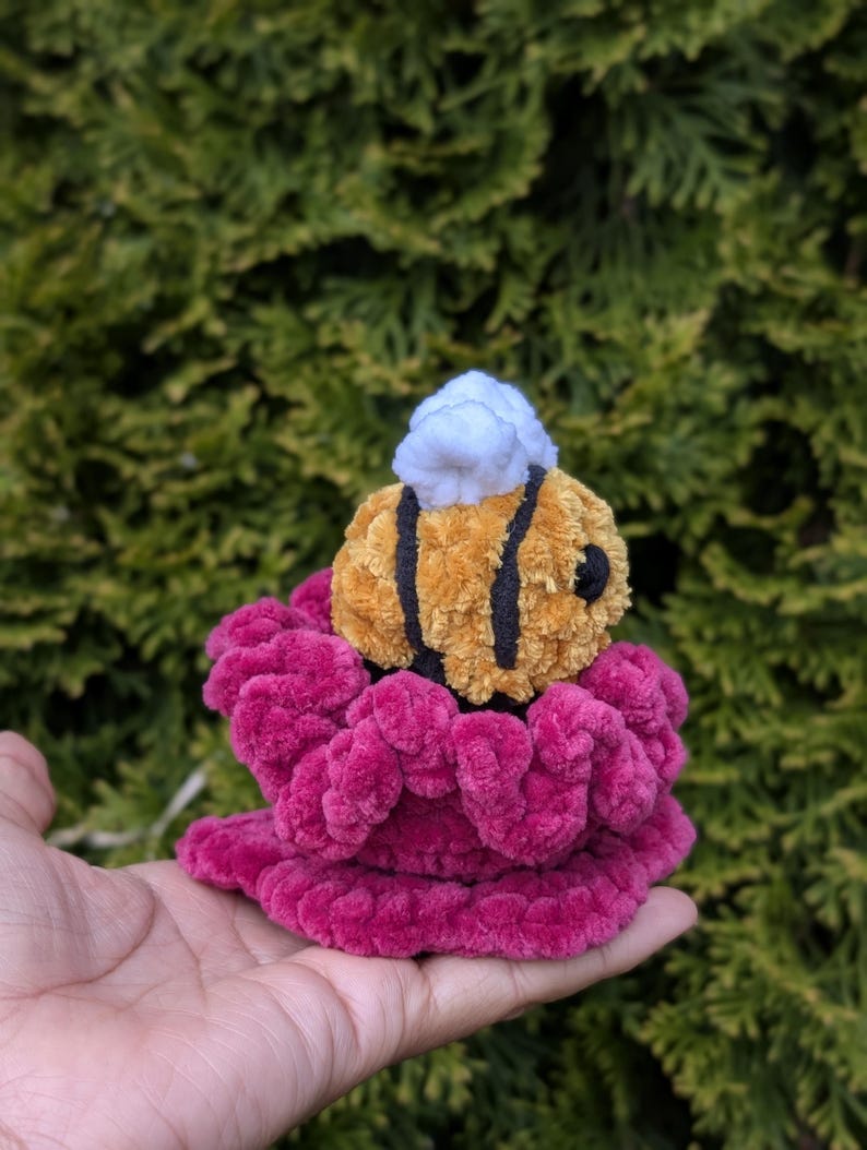 May include: A handmade plush bee with black stripes and a white head, perched atop a vibrant magenta flower. The bee is made of soft, textured material, and the flower has a ruffled appearance. The background is a blurred green.