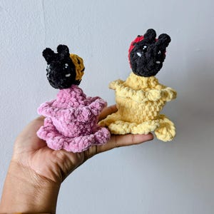 May include: Two handmade crochet dolls. One doll is pink, the other is yellow. Both dolls have black heads with white eyes and small, rounded ears. The pink doll has a yellow and black head covering, while the yellow doll has a red and black head covering.