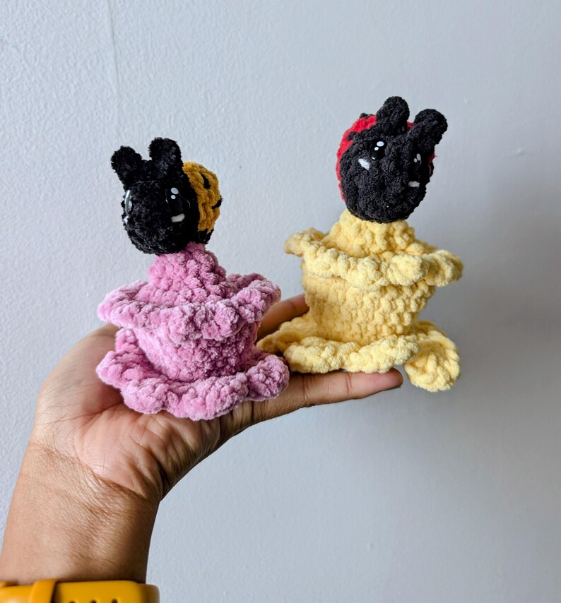 May include: Two handmade crochet dolls. One doll is pink, the other is yellow. Both dolls have black heads with white eyes and small, rounded ears. The pink doll has a yellow and black head covering, while the yellow doll has a red and black head covering.