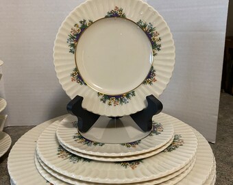 May include: A set of eight white ceramic dinner plates with a scalloped edge and a floral design in shades of purple, blue, green, and orange. The plates are stacked in a pyramid shape.