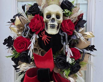 May include: A Halloween wreath featuring a skull wearing a gold crown, surrounded by red and black roses, a black velvet bow, and gold accents. The wreath is decorated with a black spider and chains.