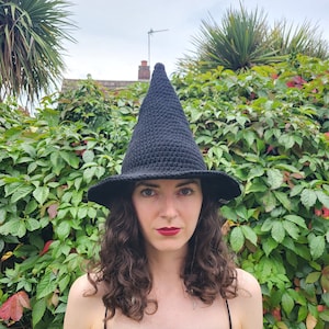 Puede incluir: Un sombrero de bruja negro de ganchillo con ala ancha y corona puntiaguda. La persona lleva el sombrero delante de un exuberante fondo verde. La persona lleva un vestido negro.