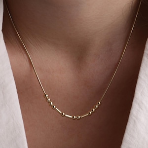 May include: A delicate gold necklace with a series of small beads and bars, creating a minimalist design. The necklace is worn against a white top and blazer, with a hand gently touching the blazer.