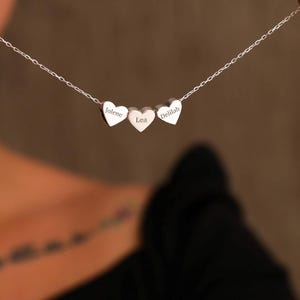 Könnte beinhalten: Eine silberne Halskette mit drei herzförmigen Anhängern. Jeder Herzanhänger trägt einen eingravierten Namen: Jolene, Lea und Delilah. Die Halskette wird vor einem unscharfen Hintergrund präsentiert.