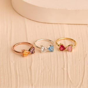 May include: Three rings featuring heart-shaped gemstones arranged in a bow-like design. The first ring is gold-toned with orange and purple stones, the second is silver-toned with clear stones, and the third is gold-toned with red and pink stones.