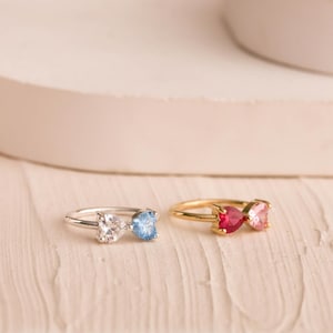 May include: Two rings with heart-shaped gemstones. One ring has a silver band with a clear and light blue stone. The other ring has a gold band with a red and pink stone. The rings are displayed on a light-colored surface.