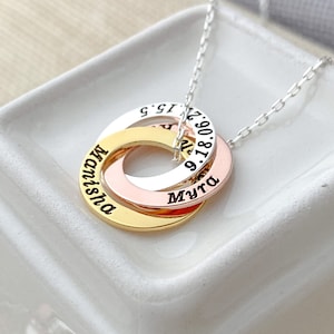 May include: A silver necklace featuring three interlocking rings in gold, rose gold, and silver. Each ring is engraved with a name and a date. The necklace is displayed on a white surface.