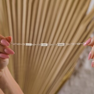 May include: A silver chain bracelet with three rectangular name tags. The tags are engraved with the names "Ashley", "Robin", and "Daniel".