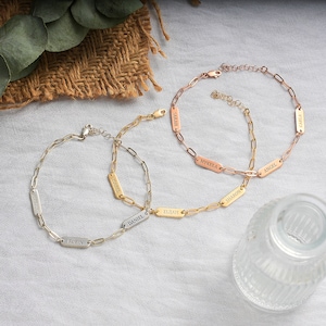 May include: Three gold chain bracelets with personalized name tags. The bracelets are on a light beige surface. The names on the tags are "Sarah", "Mykela", "Angel", "Sharon", "Elijah", "Daniel", "Isabel", "Robin", and "Carla".