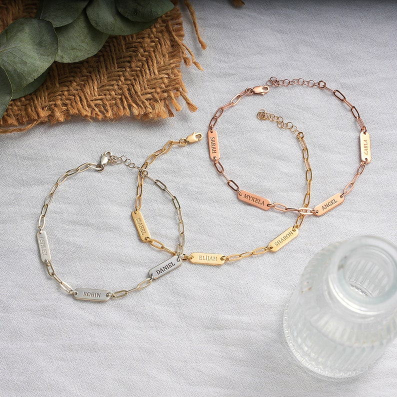 May include: Three gold chain bracelets with personalized name tags. The bracelets are on a light beige surface. The names on the tags are "Sarah", "Mykela", "Angel", "Sharon", "Elijah", "Daniel", "Isabel", "Robin", and "Carla".