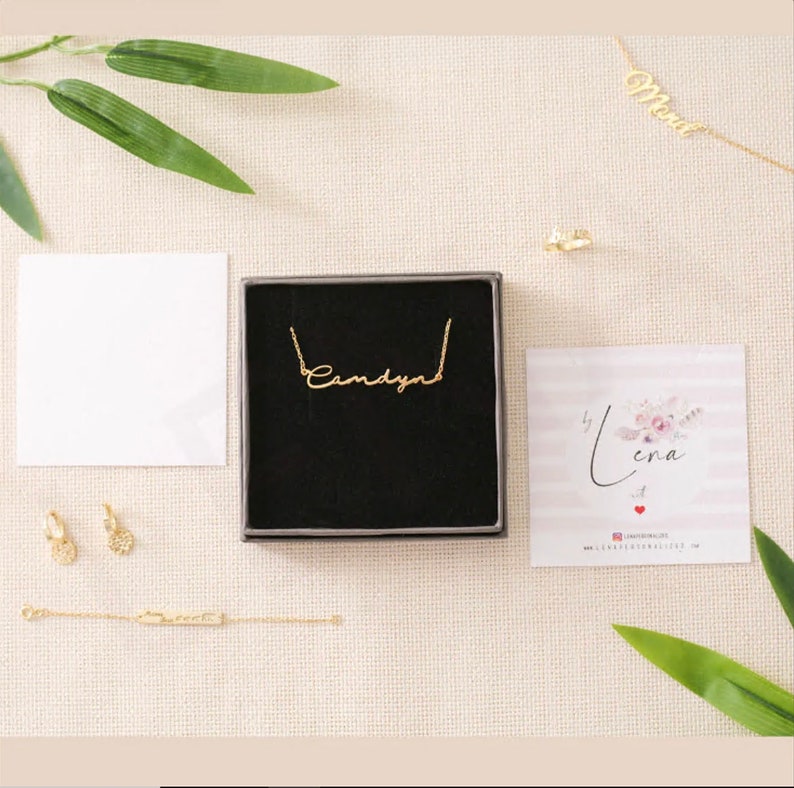 May include: A gold necklace with the name "Candyn" written in cursive script. The necklace is in a black box with a white background. There are other gold jewelry pieces in the image, including earrings and a bracelet.