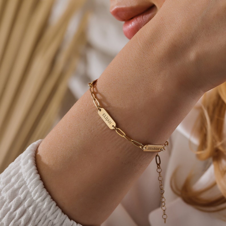 May include: Gold chain bracelet with two rectangular name tags. The name tags are engraved with the names "Elijah" and "Sharon".