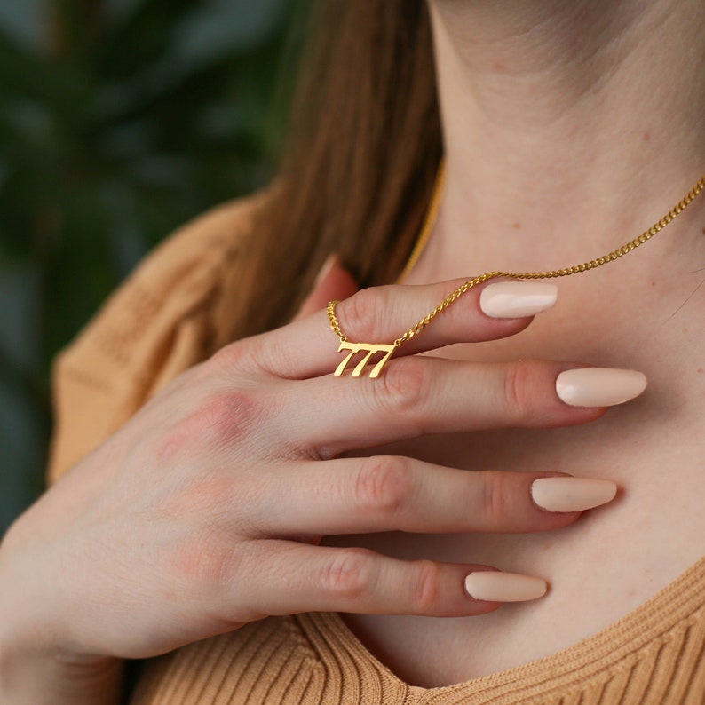 May include: A gold necklace with a pendant in the shape of the number 777. The necklace is worn by a person with light skin and long, light brown hair. The person is wearing a light brown sweater.