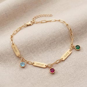 May include: Gold-colored bracelet with three name tags that read "MARY", "JOSIAH", and "ALICE". Each tag has a different colored gemstone: blue, red, and green. The bracelet is on a neutral background.