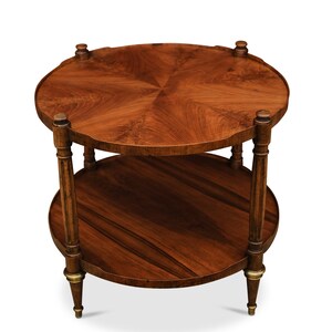 May include: A two-tiered round wooden table with a dark brown finish. The table has a scalloped edge and a decorative design on the top tier. The legs are turned and have a brass finish.