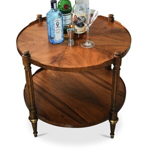 May include: A two-tiered wooden serving cart with a round top and bottom shelf. The cart has four legs and is made of dark wood with a natural wood grain. There are four bottles of liquor on the top shelf, including Bombay Sapphire gin, Tanqueray gin, Captain Morgan spiced rum, and a bottle of vodka. There is also a martini glass and a small metal jigger on the top shelf.