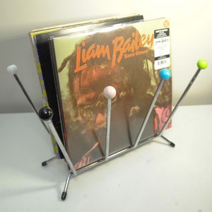 Pode incluir: Um suporte de discos de vinil cromado com cinco pontas em forma de bola coloridas segura uma pilha de discos de vinil. O disco superior é um álbum de Liam Bailey intitulado "Zero Grace".