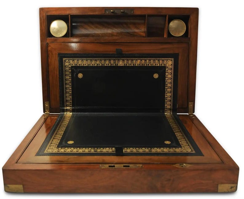 May include: A wooden writing desk with a leather writing surface and gold accents. The desk is open and the interior is visible. The desk has a hinged lid and a small drawer.