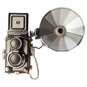 Puede incluir: Una cámara réflex de doble lente Yashica-44 vintage con una unidad de flash conectada. La cámara es negra y plateada con el nombre "Yashica-44" impreso en la parte delantera. El flash tiene un reflector circular grande.