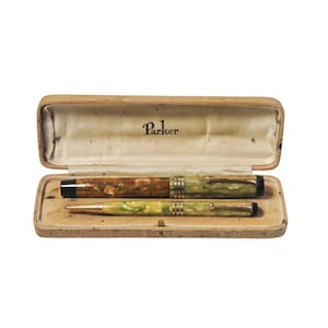 Puede incluir: Un juego de bolígrafo y lápiz vintage Parker en una caja marrón. El bolígrafo y el lápiz son ambos de color verde y marrón veteado con detalles dorados. La caja tiene la palabra "Parker" impresa en el interior.
