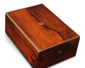 Op de afbeelding: Een rechthoekige houten kist met een rijke, roodbruine afwerking en messing afwerking. Het deksel heeft een opvallend houtnerfpatroon en een kleine, ovale metalen accent. Een sleutelgat is zichtbaar aan de voorkant.
