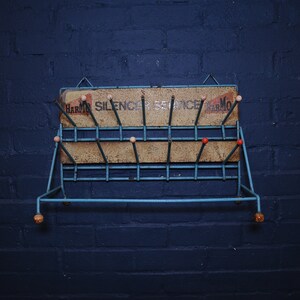 May include: A blue metal shoe rack with a sign that reads "Harmon Silencer Service Harmon". The rack has wooden knobs and is mounted on a blue brick wall.