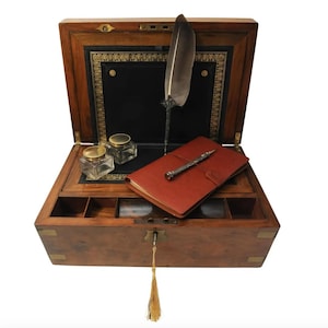 May include: An antique wooden writing box with a leather interior, a quill pen, a small notebook, and two inkwells. The box has brass accents and a keyhole.