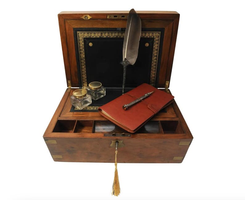 May include: An antique wooden writing box with a leather interior, a quill pen, a small notebook, and two inkwells. The box has brass accents and a keyhole.