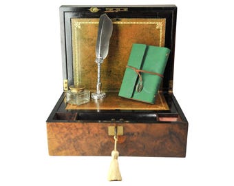 Op de afbeelding: Een houten doos met een wortelhouten afwerking en een sleutel met een kwastje, met daarin een zilveren ganzenveer, een doorzichtige glazen inktpot met een gouden deksel en een groen lederen dagboek vastgezet met een bruin leren koord en een metalen blaadje.