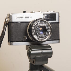 Op de afbeelding: Een Olympus Trip 35 camera, zilver en zwart, gemonteerd op een zwart statief. De camera heeft een prominente lens en de tekst "OLYMPUS TRIP 35" is zichtbaar. Een zwarte polsriem is aan de camera bevestigd.