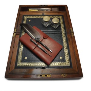 May include: A wooden writing desk with a leather writing surface. The desk is open and has a red leather journal, two inkwells, a quill pen, and two metal pens on the surface.